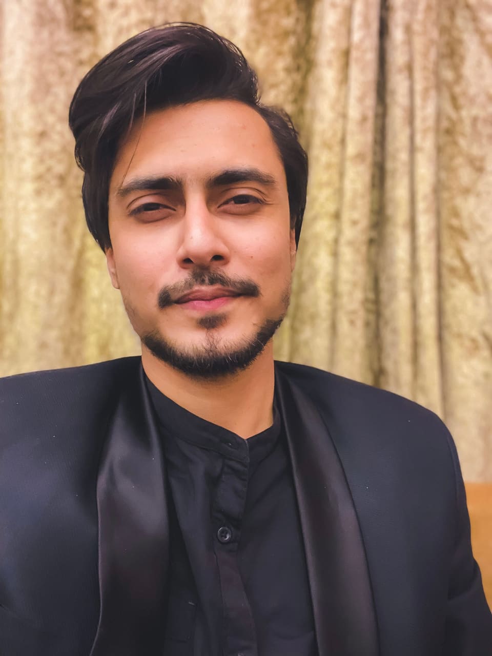 Faizaan Salar, founder of Fana Studios, in a black bandhgala in front of a gold velvet curtain