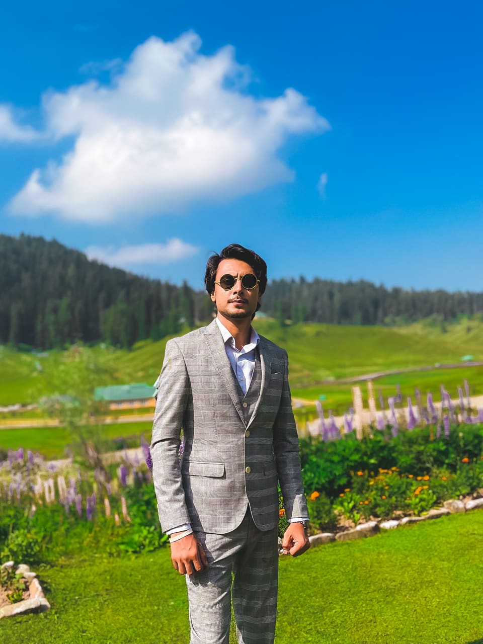 Faizaan Salar in a grey check suit and round sunglasses, standing in a meadow in Kashmir